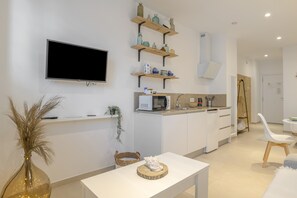A modern kitchen with white cabinets, a flat-screen TV, wooden shelves with kitchenware, a dining area with a white table and chairs, and a small plant. - Comfortable "Tarifa Dreams" Studio, Excellent Location. Perfect for Couples (Tarifa)