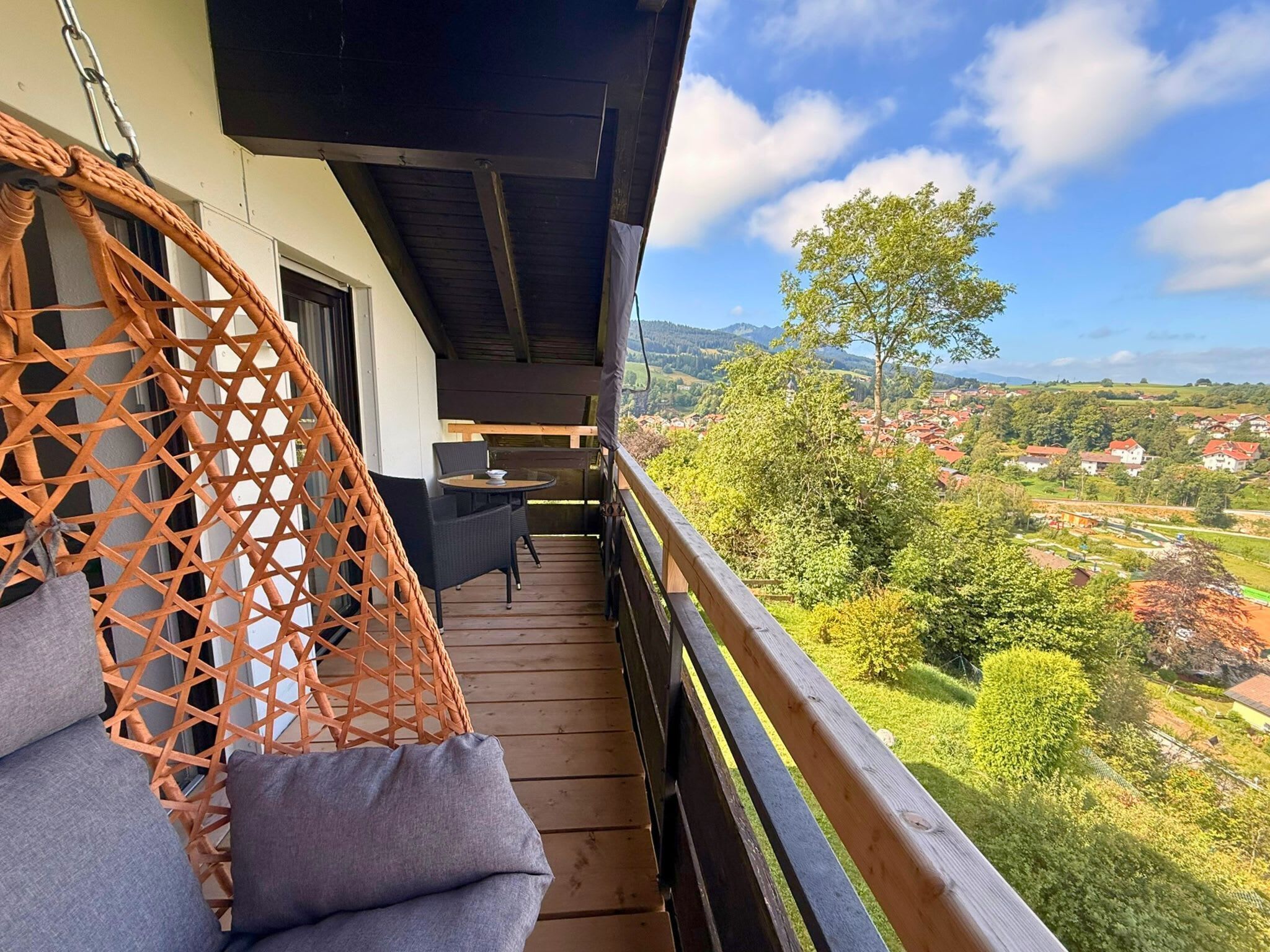 Eine Holzterrasse mit Korbstuhl und Tisch, mit Blick auf ein Dorf und Berge.