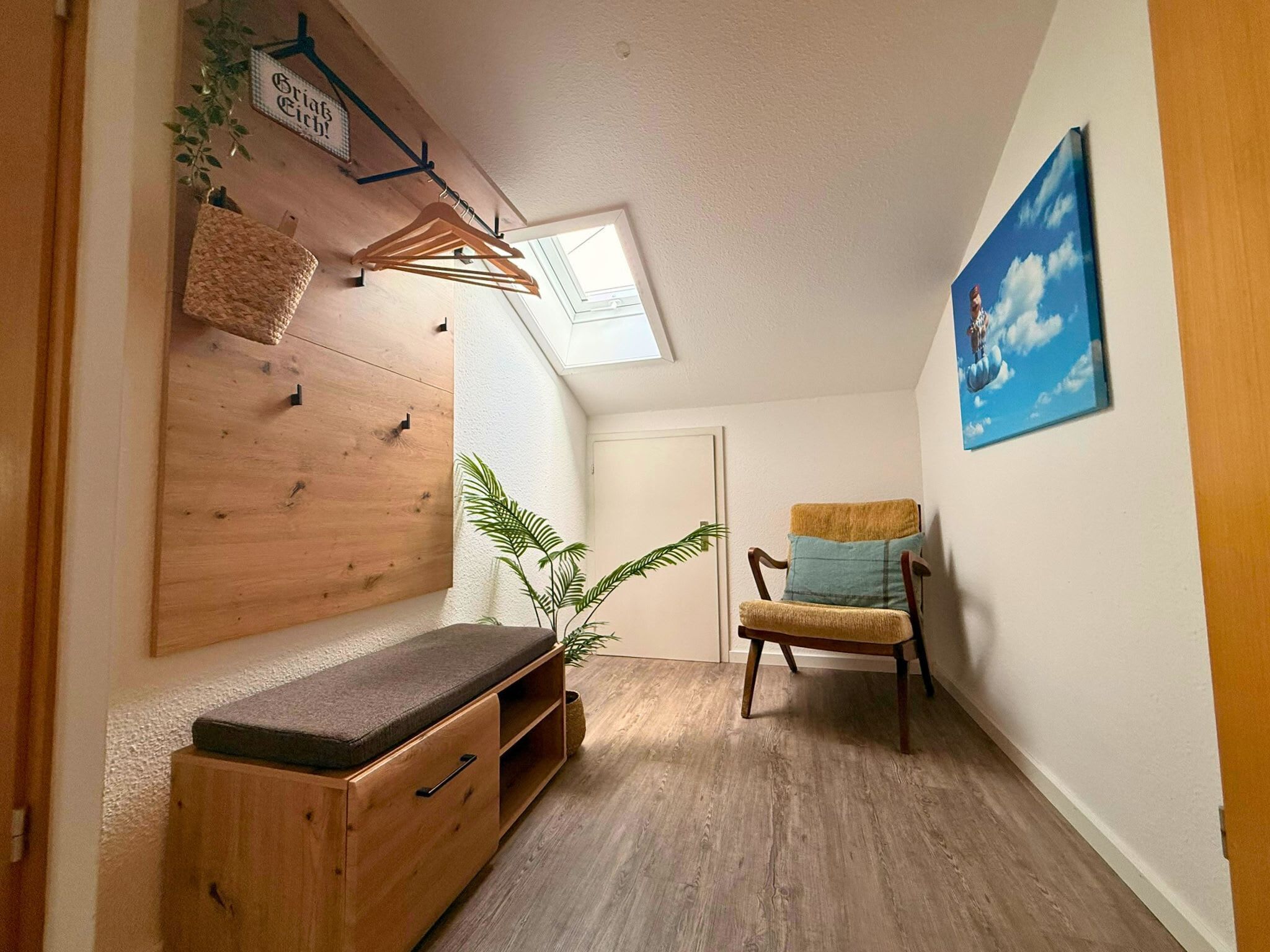 Un couloir étroit avec un banc en bois, une chaise avec des coussins, une plante suspendue et un tableau encadré au mur.