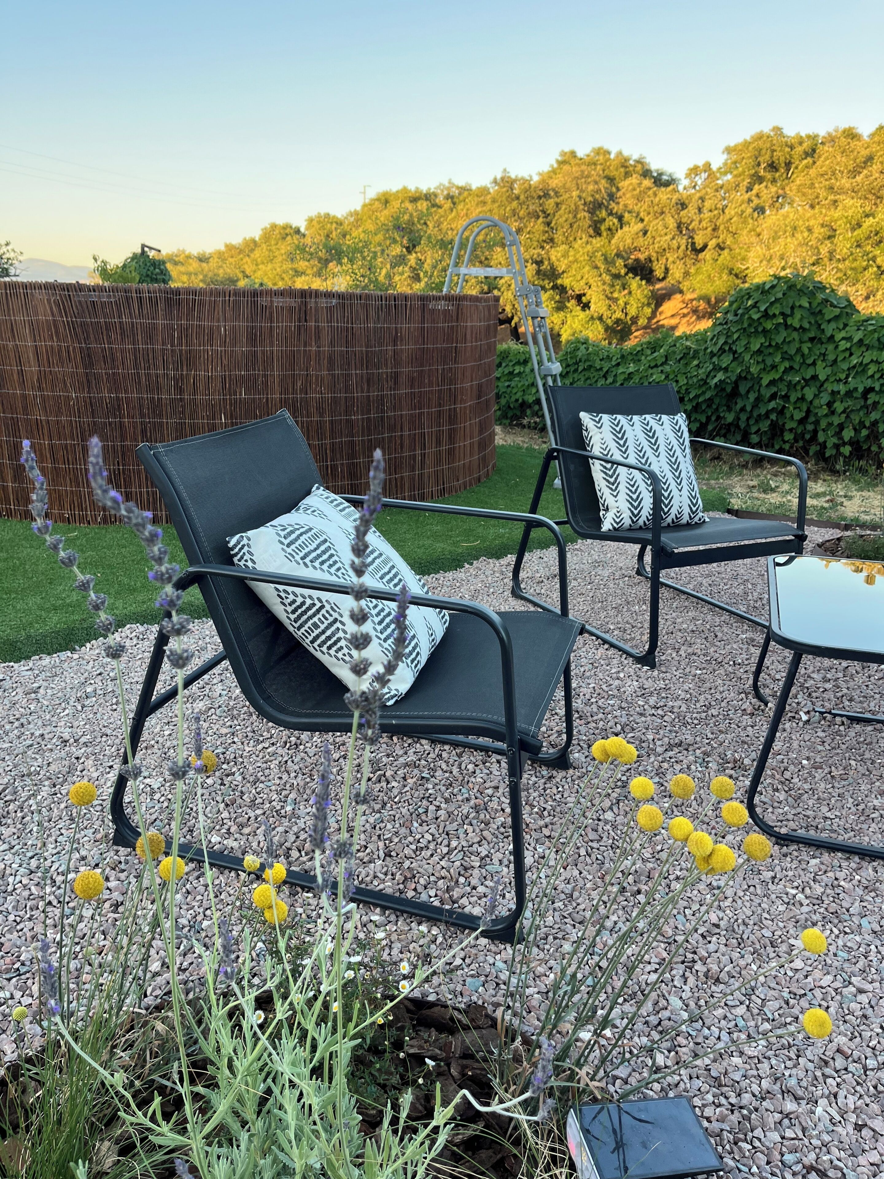Dos sillas de metal negro con cojines estampados, una mesita y una escalera en un jardín con suelo de grava y follaje.