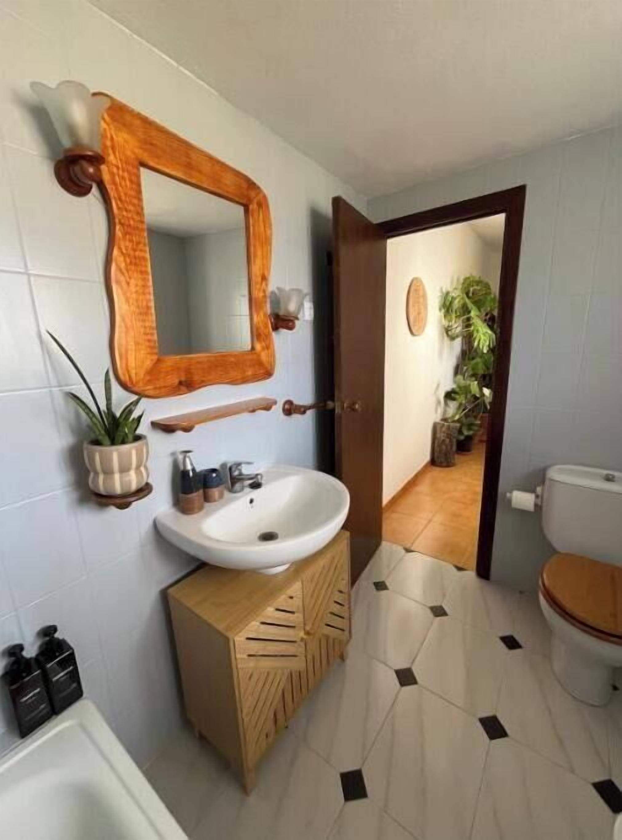 Un baño con tocador de madera, un lavamanos blanco y un espejo. Hay una planta en maceta a la izquierda y un inodoro visible a través de una puerta abierta.