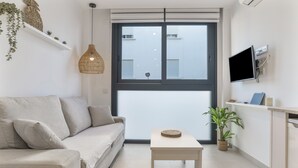 A modern living room with a sofa, a table, a hanging light fixture, a TV mounted on the wall, and a window with blinds.