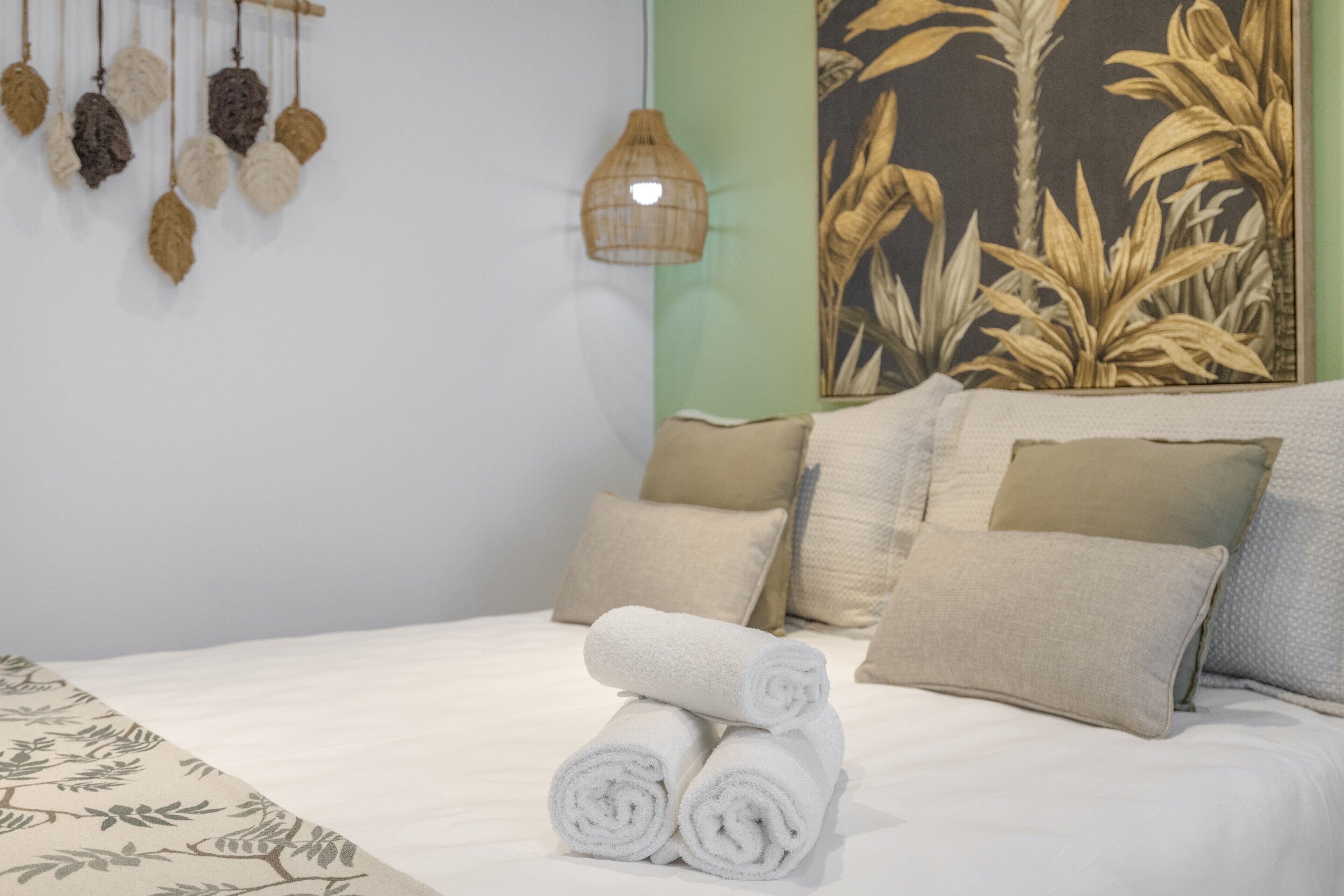 A neatly made bed with rolled white towels, a hanging plant with brown leaves, and a wall with a large botanical print.