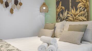 A neatly made bed with rolled white towels, a hanging plant with brown leaves, and a wall with a large botanical print.
