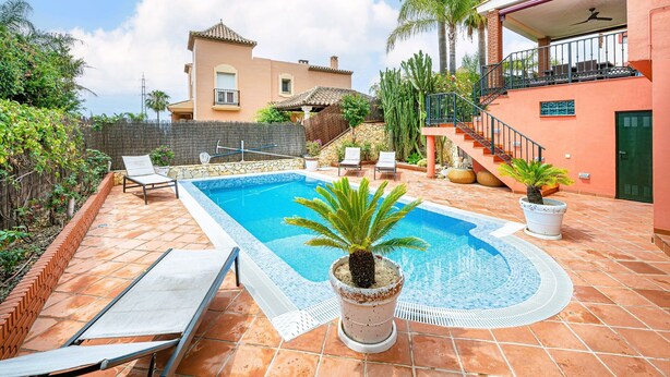 Piscina com espreguiçadeiras, um vaso de planta e um edifício com varanda ao fundo.