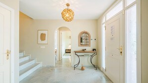 Um corredor amplo com chão de mármore, um candelabro, um aparador e uma porta com painel de vidro jateado.