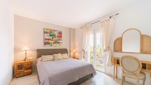 Quarto com cama, mesas de cabeceira, escrivaninha, cadeira, janela grande com cortinas e uma obra de arte na parede.