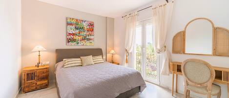 A bedroom with a bed, bedside tables, a desk, a chair, a large window with curtains, and a wall art piece.