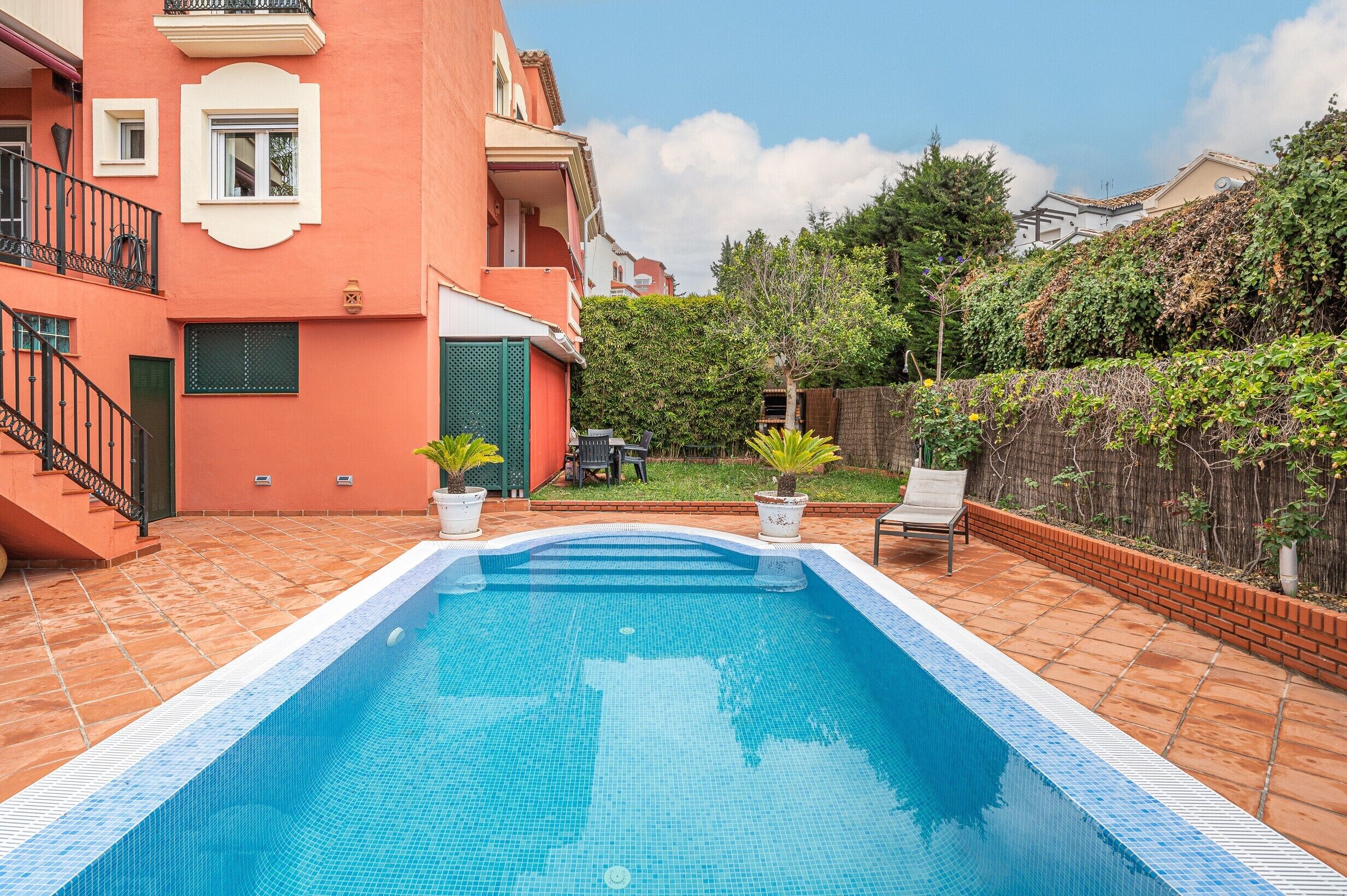 A swimming pool with tiled edges, surrounded by a brick patio and a small garden area with potted plants.