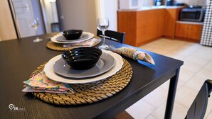 Una mesa de comedor dispuesta con platos, tazones y una copa de vino, con una cocina al fondo.