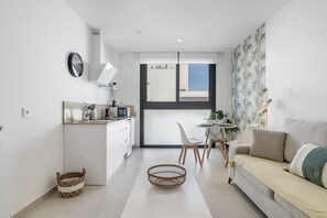 Una cocina moderna con armarios blancos, un comedor con mesa y sillas blancas, y una sala de estar con sofá y planta.