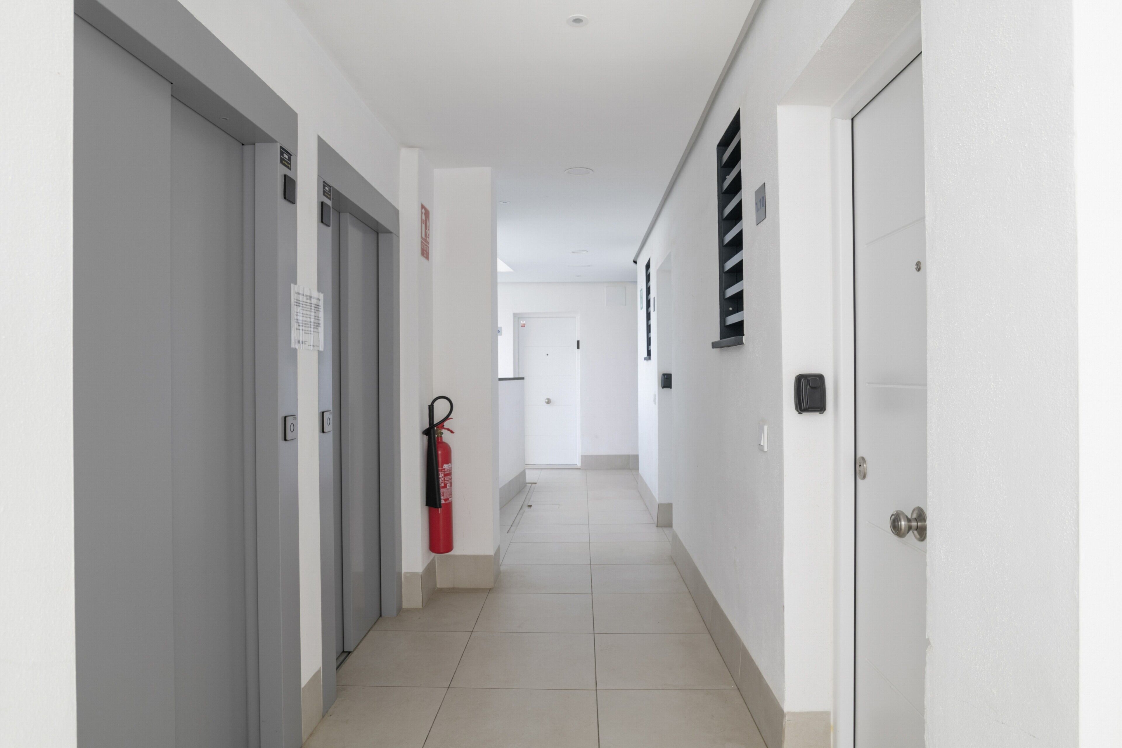 A hallway with doors, a fire extinguisher, and a vent on the wall.