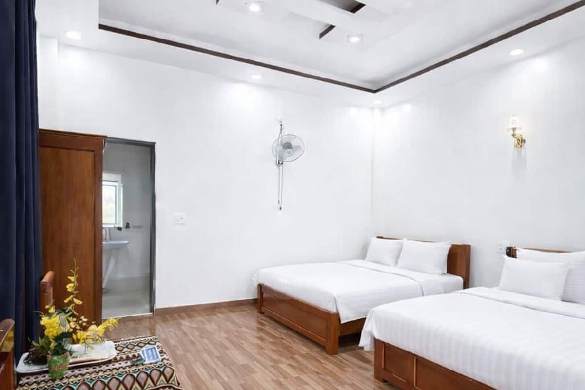 Une chambre d’hôtel avec deux lits, une table en bois avec une chaise, un ventilateur de plafond et une petite table ornée de fleurs.