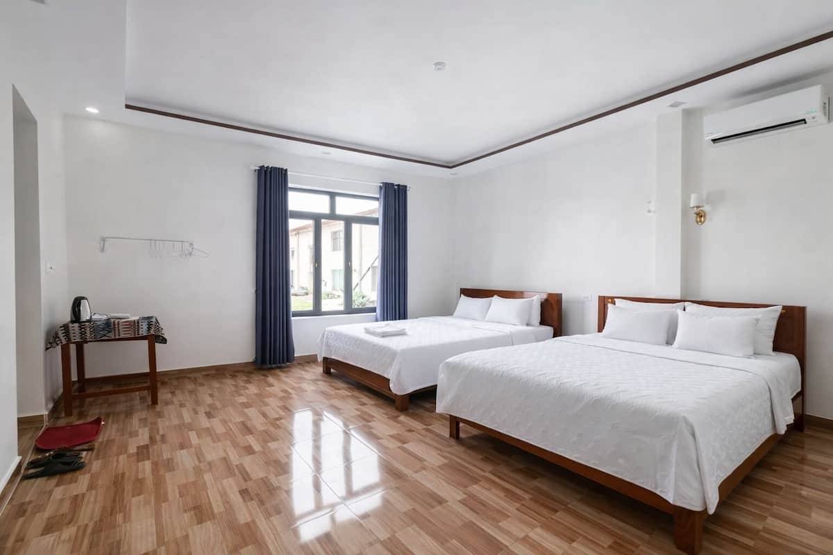 Une chambre d’hôtel avec deux lits simples, un plancher en bois, une fenêtre avec des rideaux, une petite table avec une bouilloire et un appareil de climatisation.