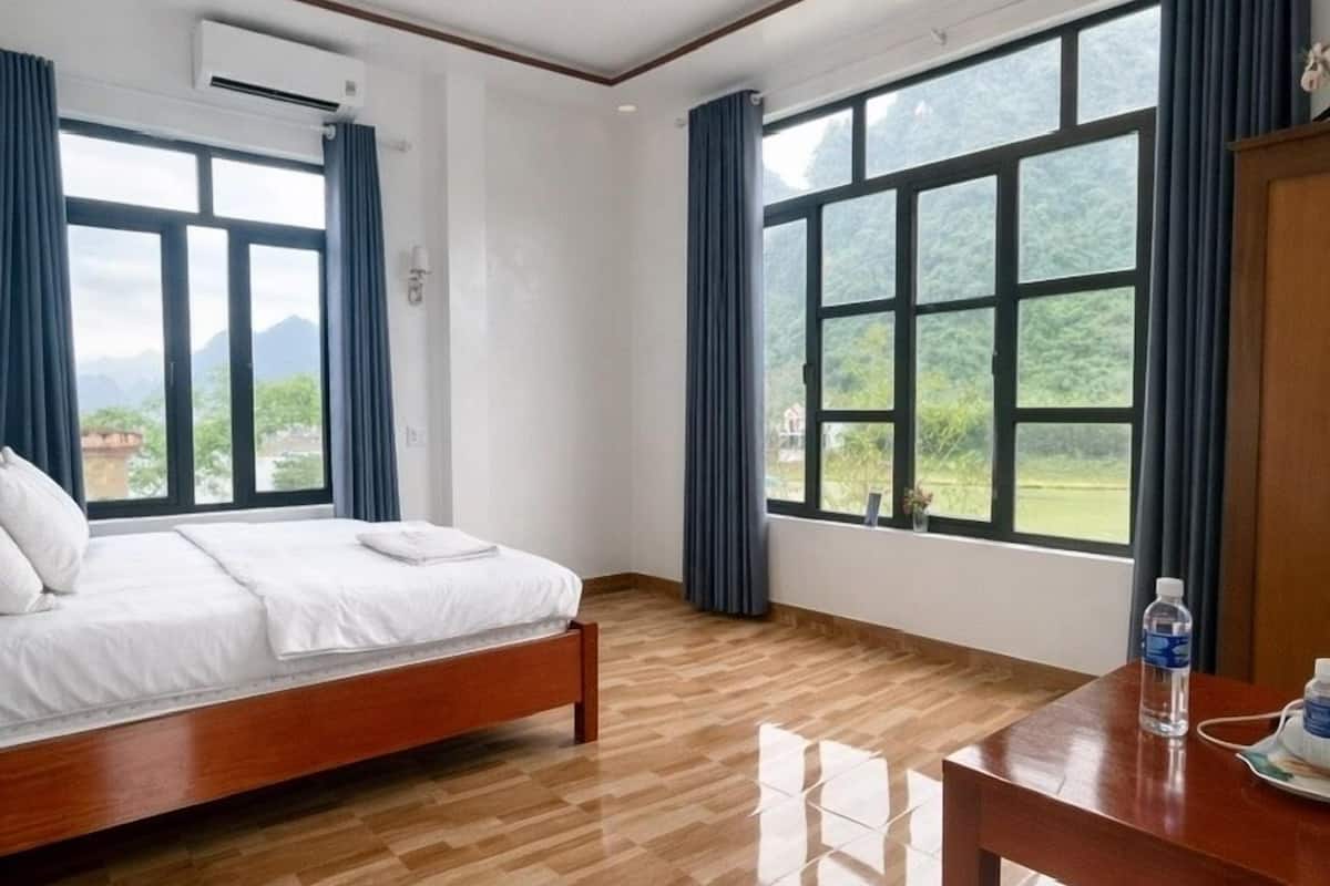 Une chambre d’hôtel avec un lit, une table en bois et de grandes fenêtres donnant sur un paysage verdoyant et montagneux.