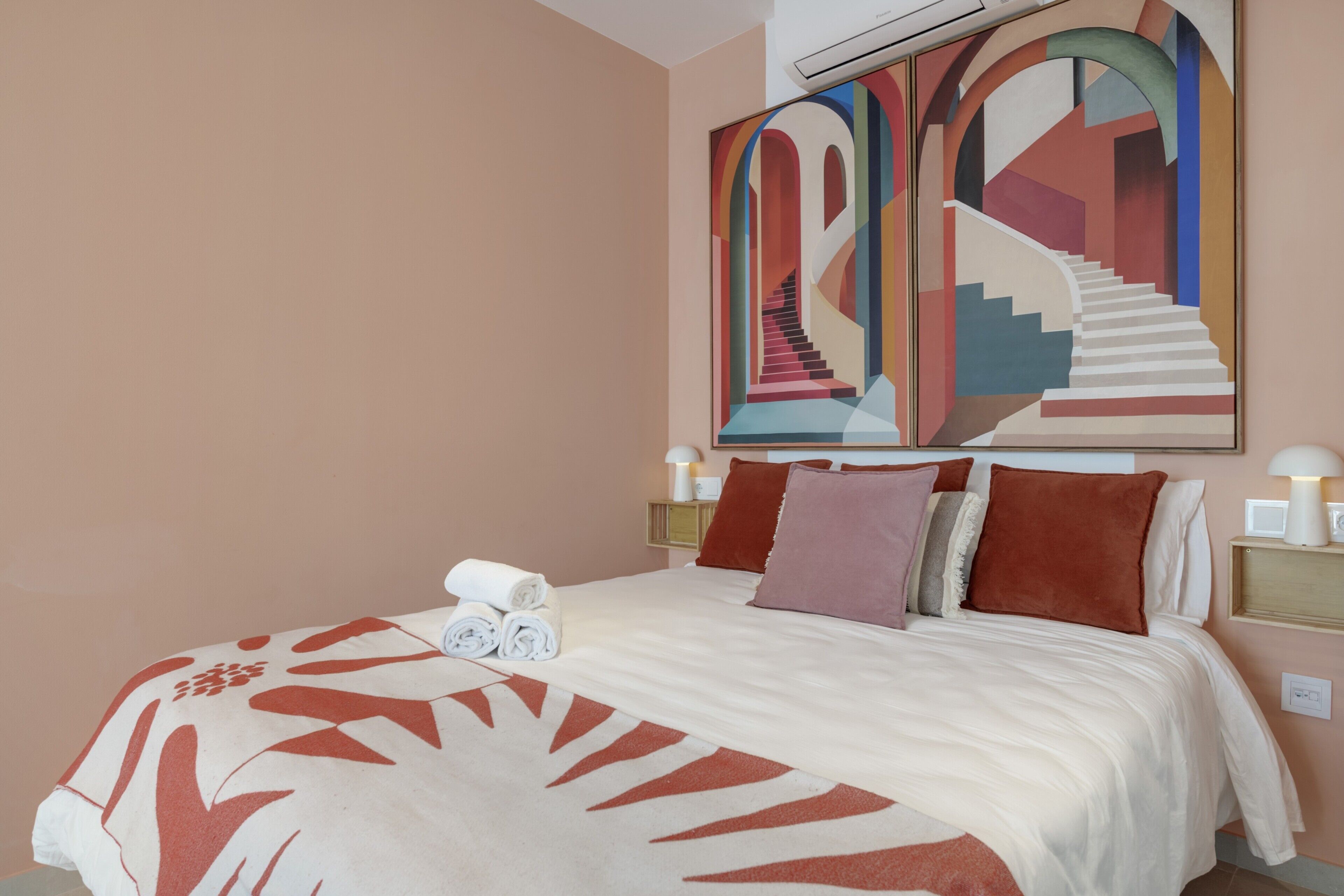 A hotel room with a bed, pillows, a blanket, a wall art piece, and a nightstand with a lamp.