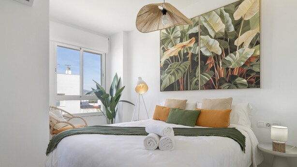 A bedroom with a bed, pillows, a bedside lamp, a large plant, and a window. - 'San Mateo Loft' Studio with Communal Pool, Wi-Fi, and Air Conditioning (Tarifa)