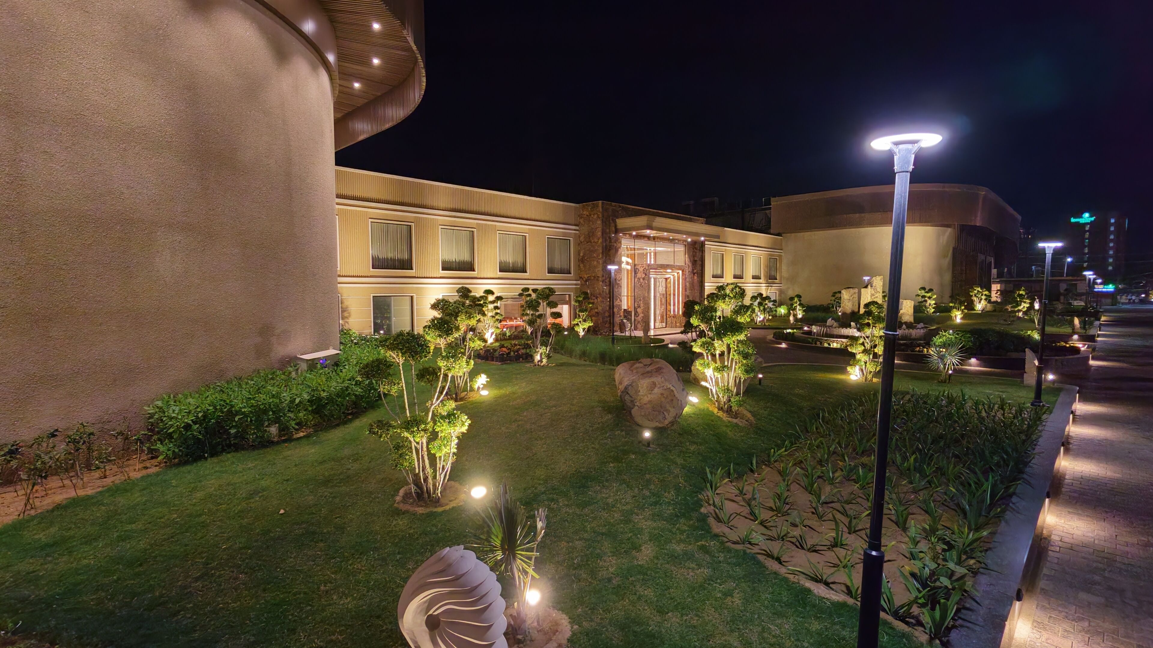 A well-lit hotel courtyard with a large building, landscaped garden, and outdoor seating area.