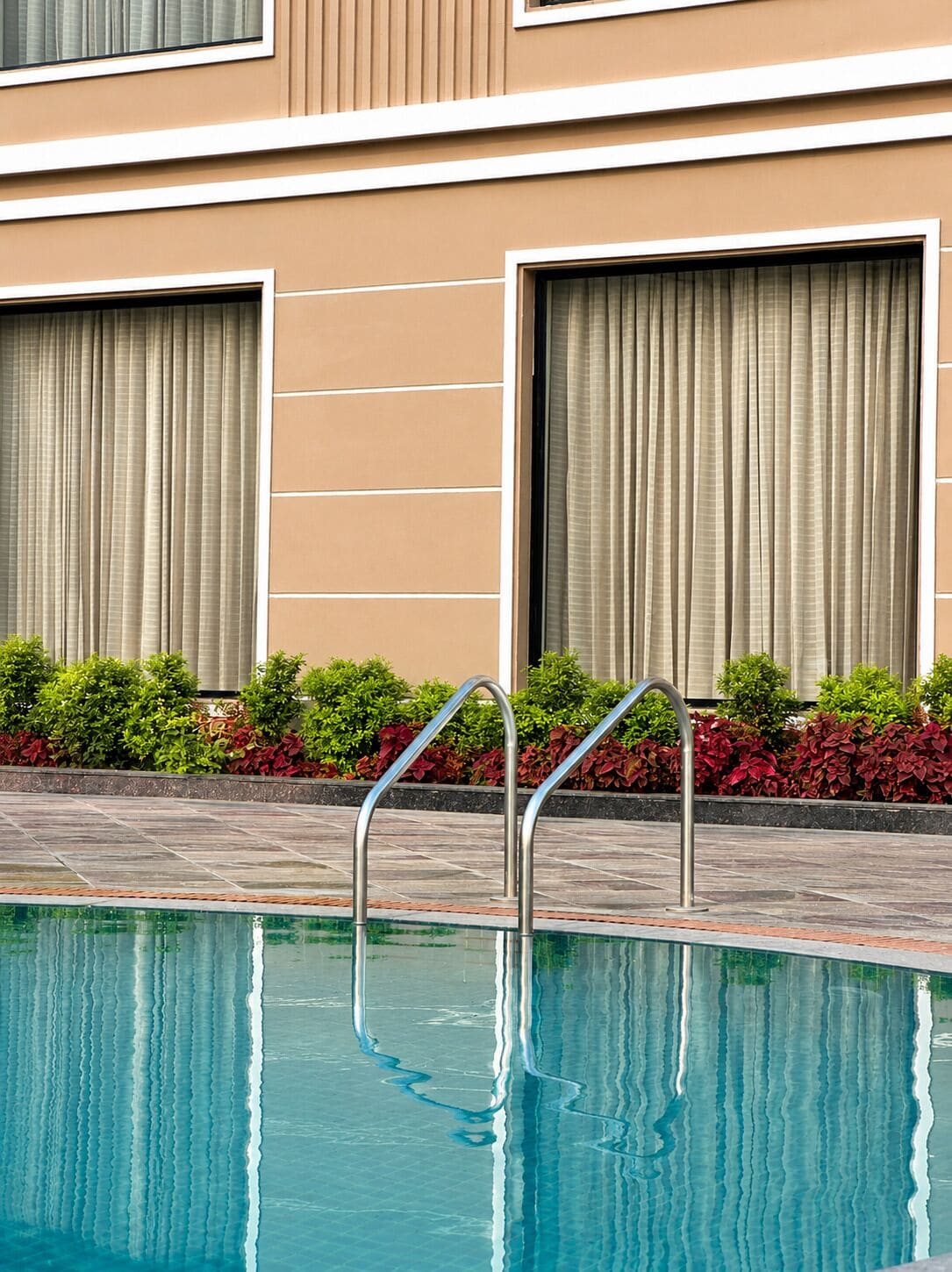 Une piscine entourée de rampes métalliques, une terrasse carrelée et un bâtiment doté de grandes fenêtres et d’une façade beige.