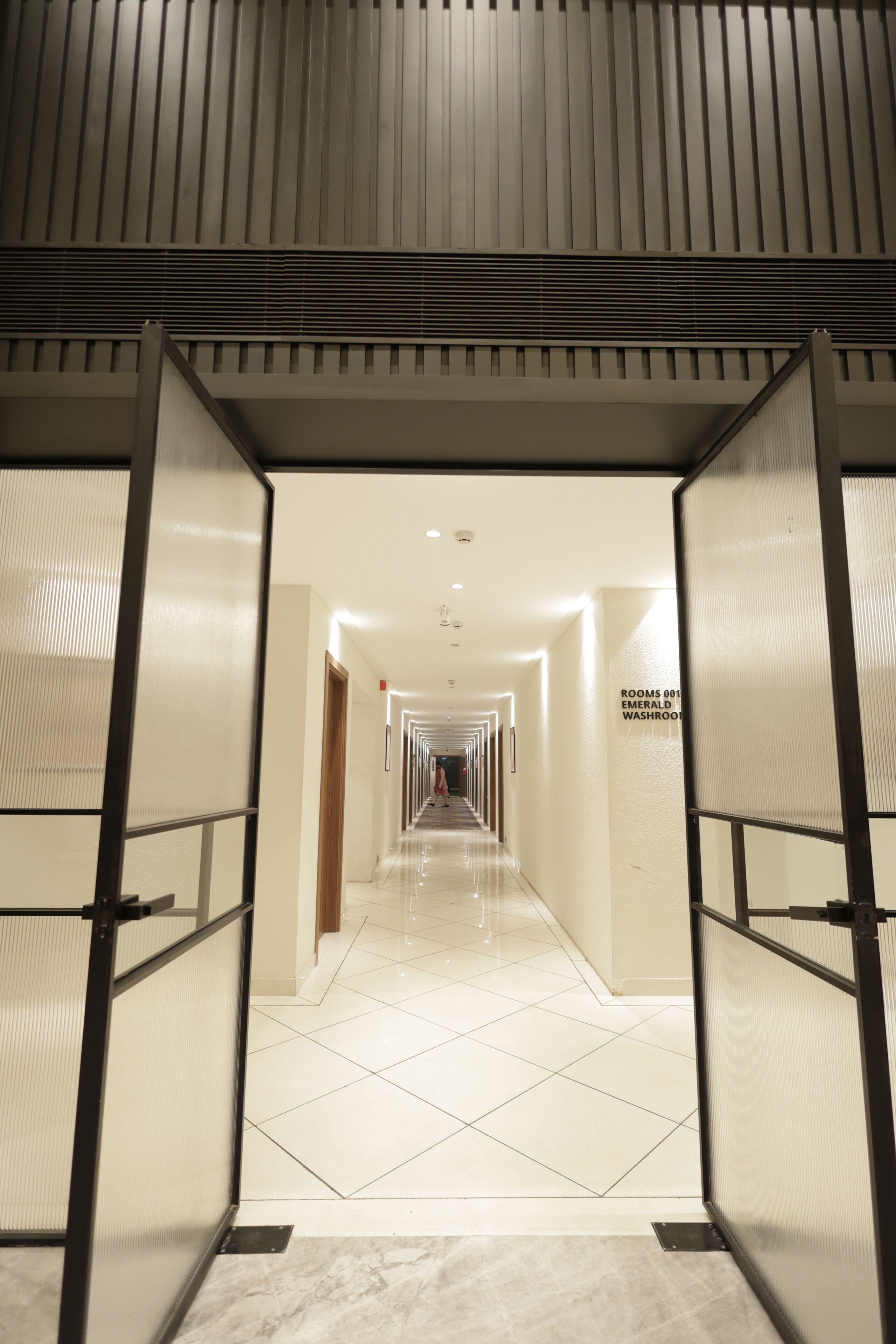 A hotel corridor with a tiled floor, a sign indicating rooms 001 to 005, and a modern design with a high ceiling and recessed lighting.