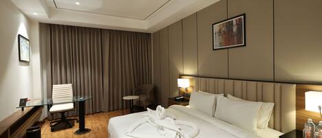 A hotel room with a large bed, a desk, a chair, a small table, a nightstand with a lamp, and a painting on the wall.