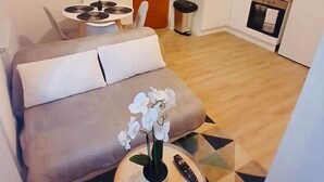 Un espace de vie compact comprenant un canapé, une petite table ronde avec une télécommande et un pot d’orchidée, une kitchenette avec une cuisinière et un évier, et une porte.