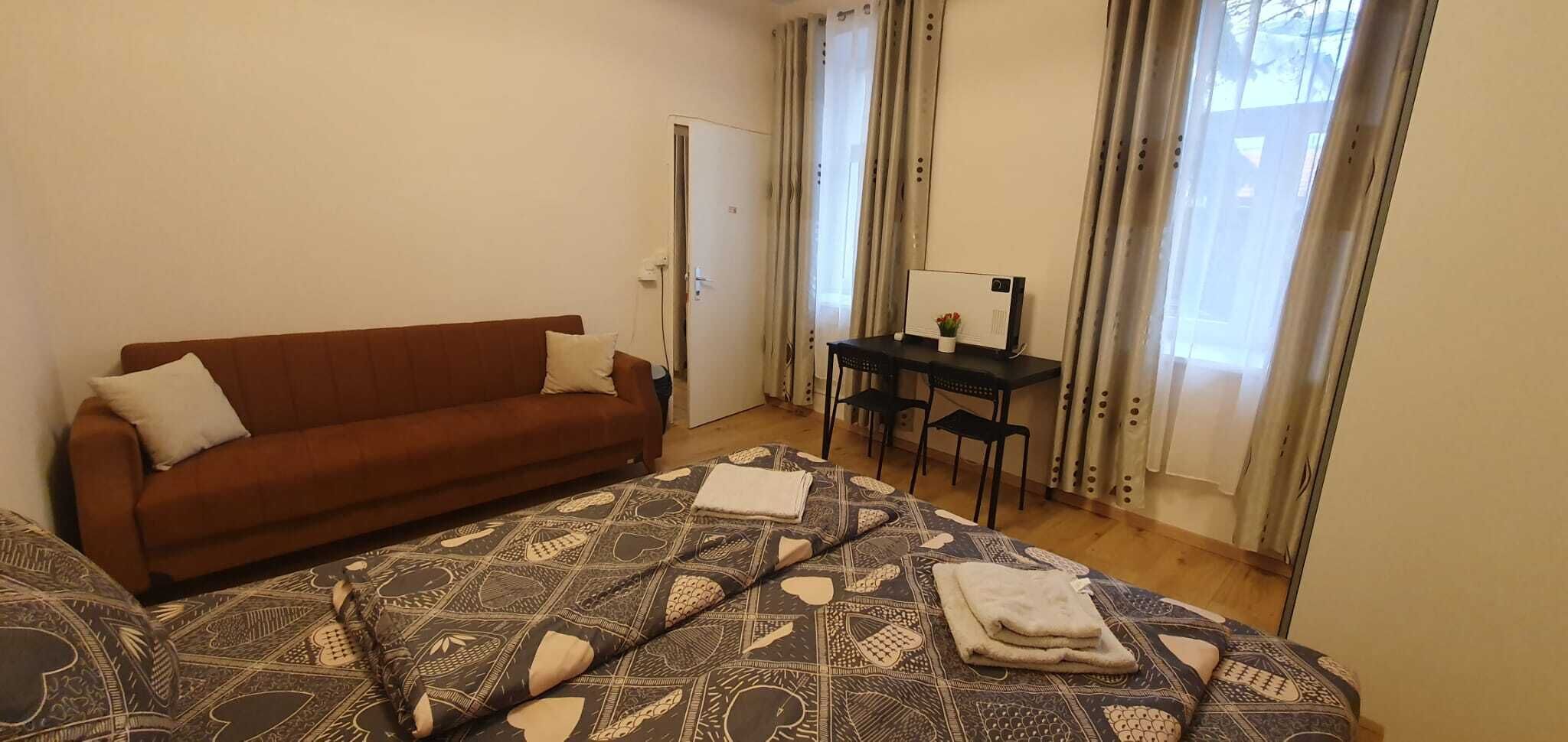 A bedroom with a brown sofa, a patterned bedspread, a desk with a chair, and a window with curtains.