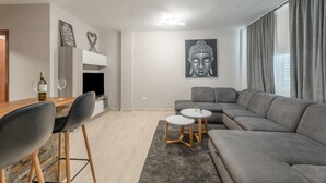 Ett modernt vardagsrum med en grå soffa, en matplats med ett träbord och stolar, en målning av Buddha och ett stort fönster med gardiner.
