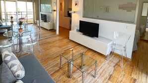 Ein modernes Wohnzimmer mit einem Glastisch, einer Couch, einem Fernseher auf einem weißen Schrank und einem großen Spiegel an der Wand.