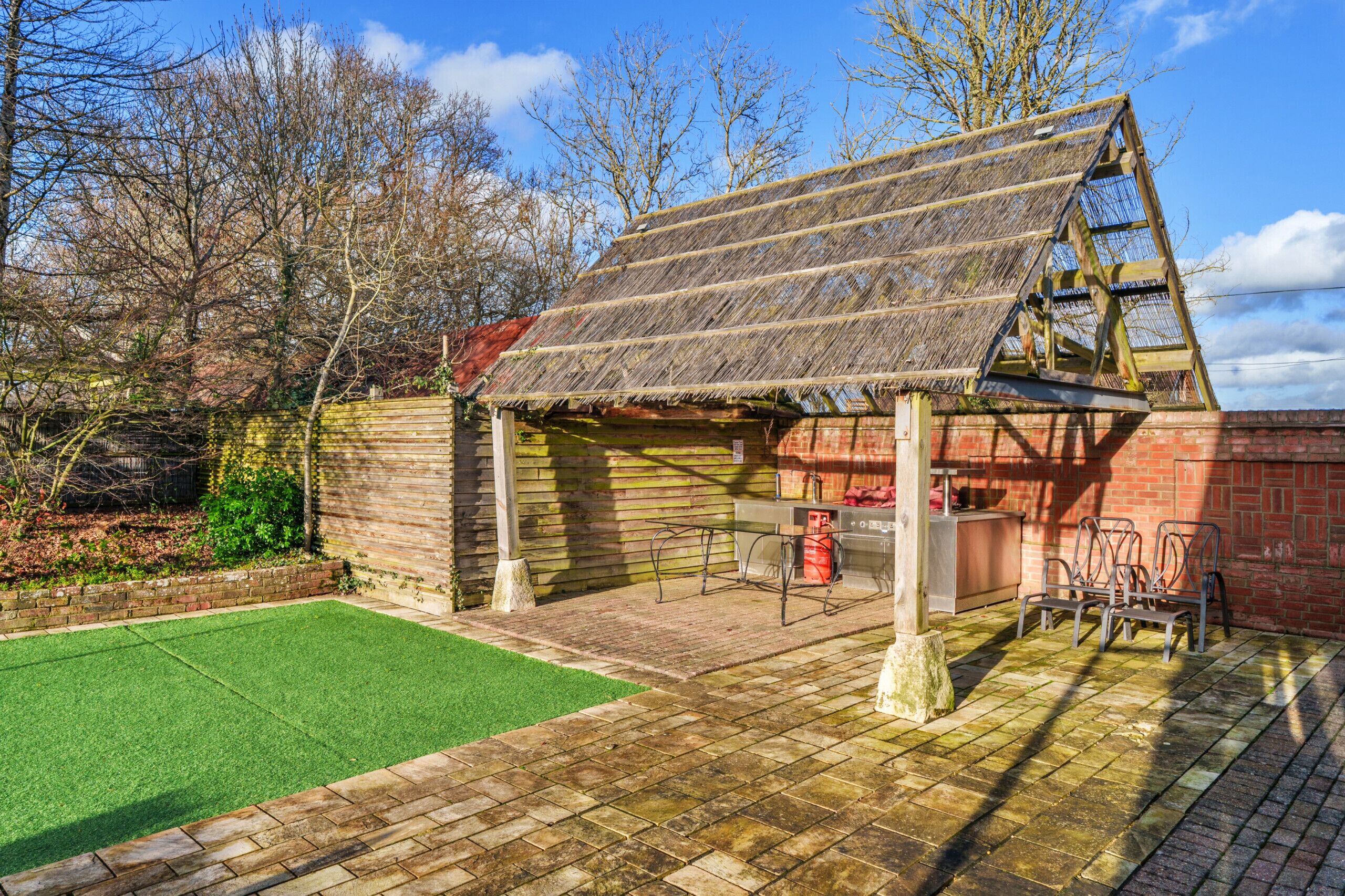 A rustic wooden structure with a sloped roof, a brick wall, and a small patio area with a green lawn and metal chairs.
