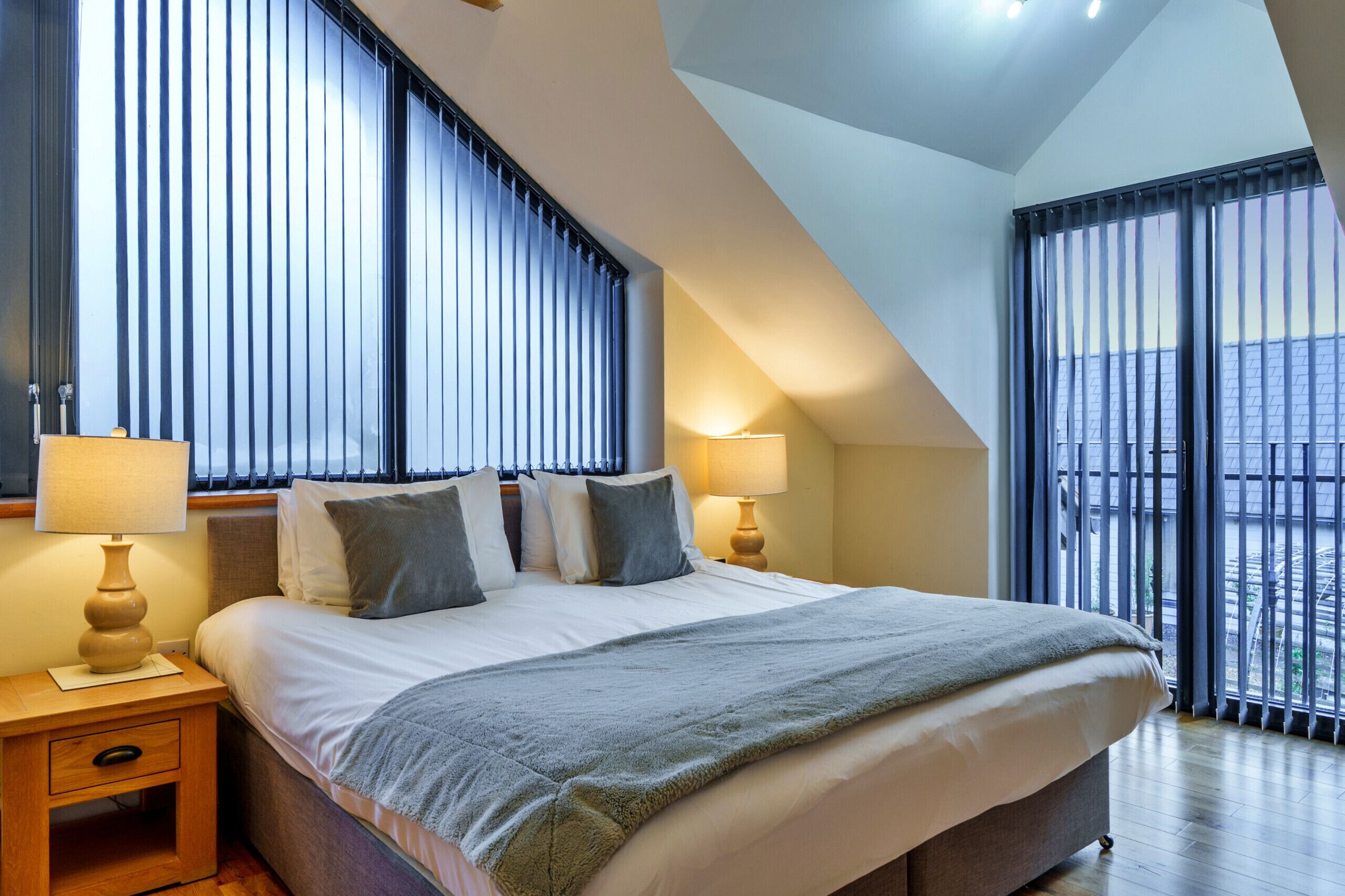 Une chambre moderne avec un grand lit, deux tables de chevet avec des lampes et une vue sur l’extérieur grâce à de grandes fenêtres du sol au plafond.