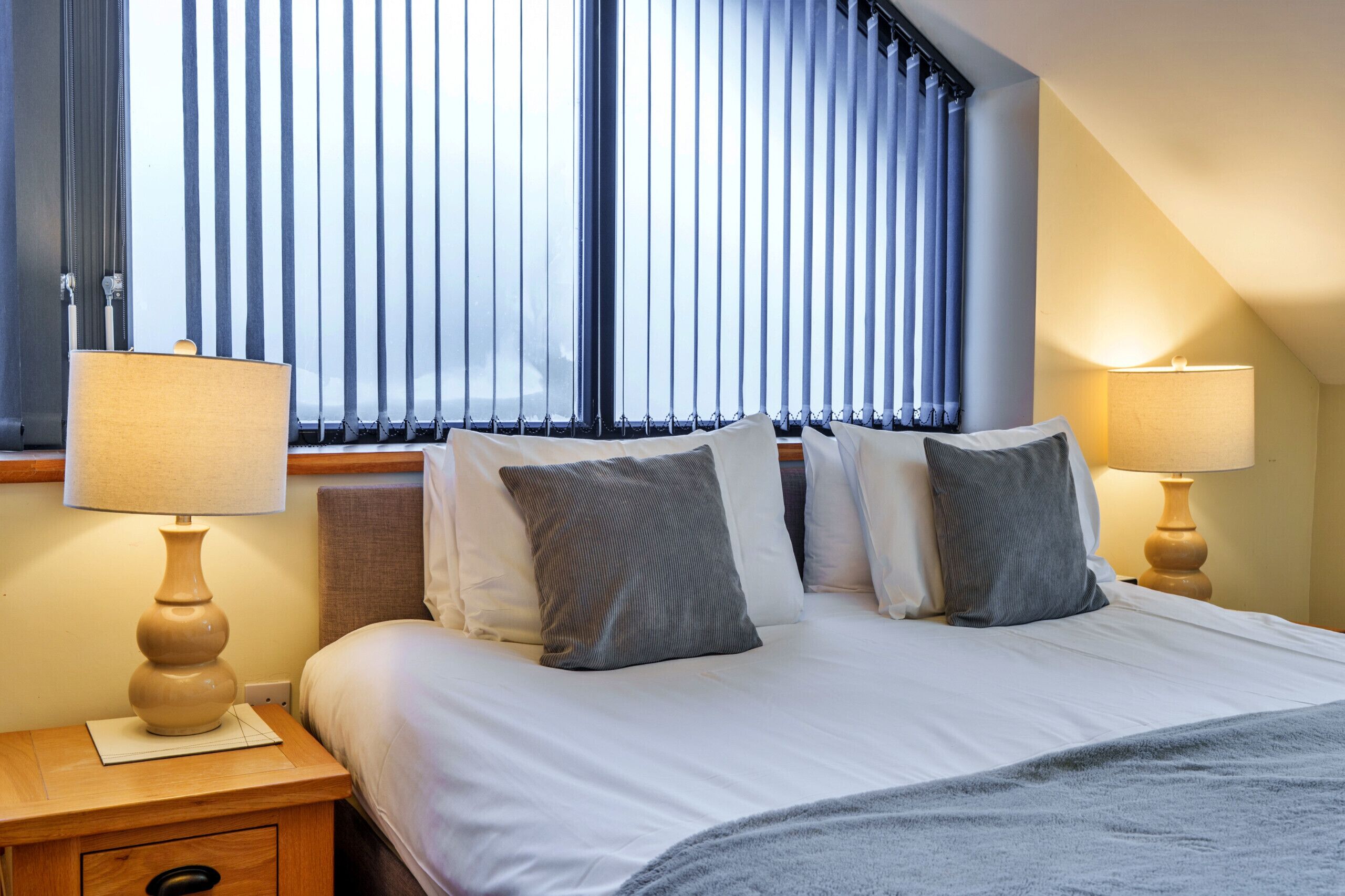 Habitación de hotel con cama, dos lámparas de noche, ventana con persianas verticales y mesita de noche con cajón.