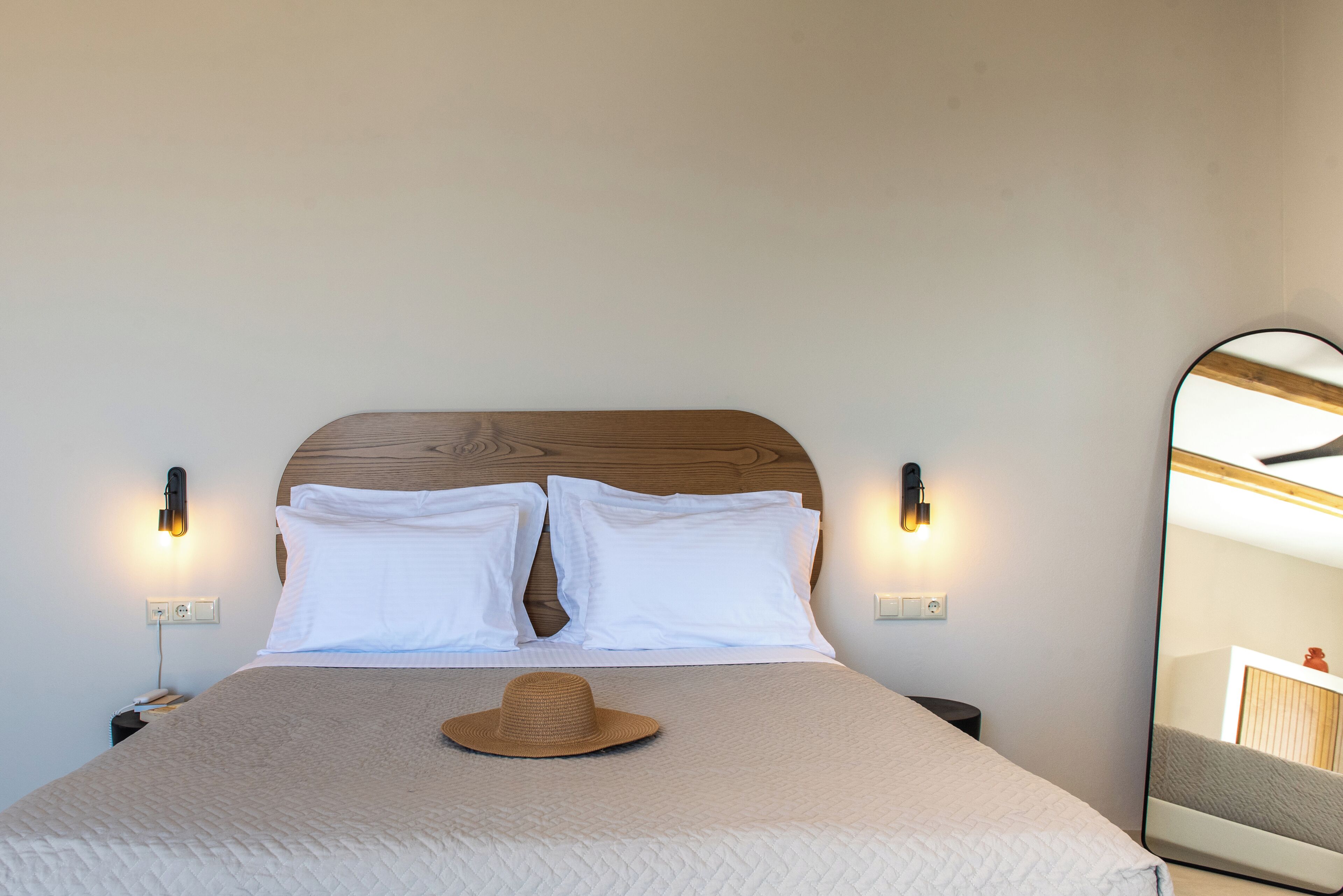 A neatly made bed with a wooden headboard, two white pillows, a beige blanket, and a straw hat on the bed. There are two wall-mounted nightstands with lamps and a large mirror on the wall.