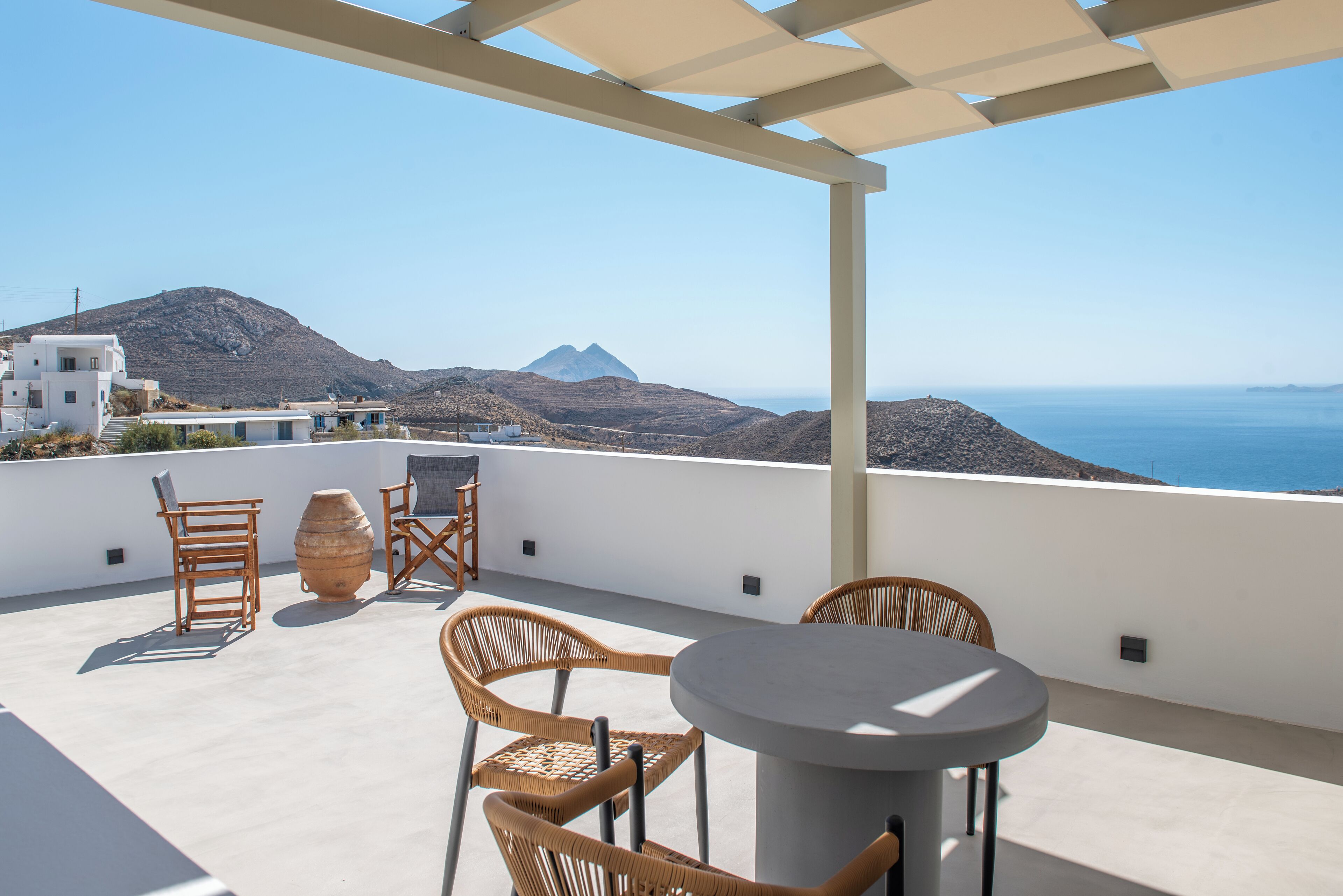 A terrace with wicker chairs and a table, overlooking a mountainous landscape and the sea.