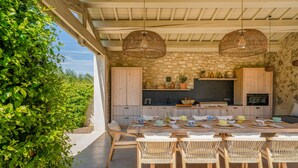 Une cuisine extérieure avec des murs en pierre, des armoires en bois et un coin repas agrémenté de chaises en osier et d’une table dressée pour le repas.