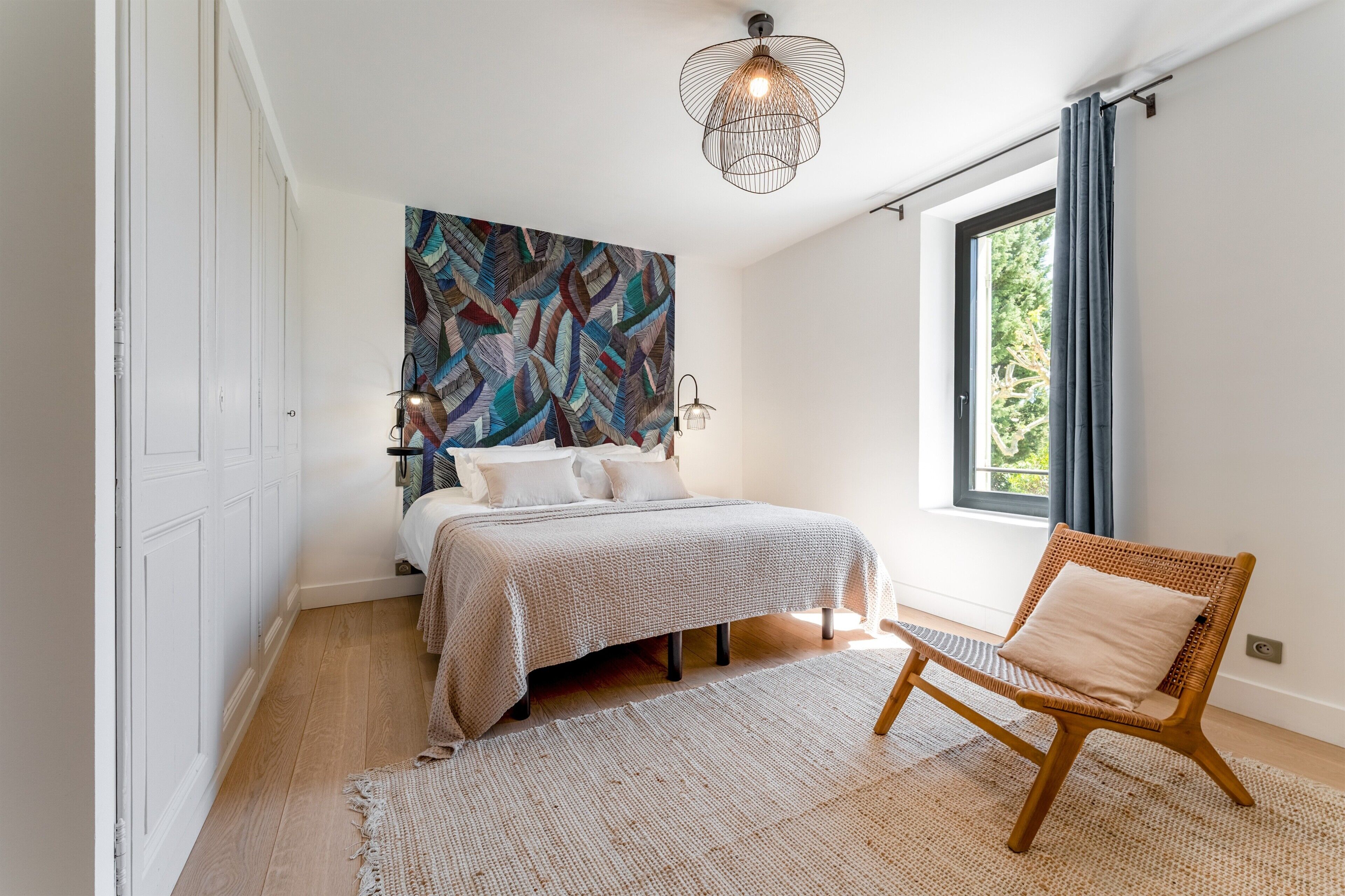 A bedroom with a bed, a chair, a large abstract painting, and a window with curtains.