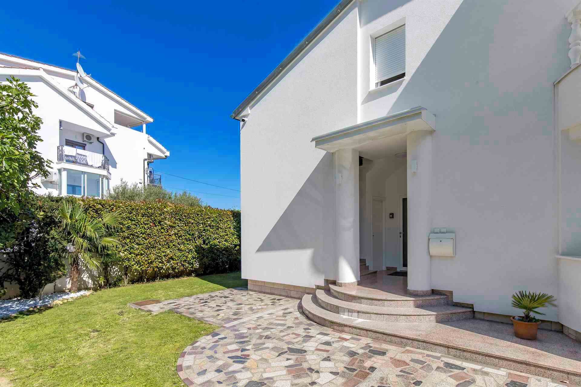 Ein modernes, zweistöckiges Haus mit weißer Fassade, einer kleinen Veranda und einem gepflasterten Einfahrtsweg zum Eingang.