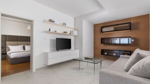 Un salon moderne doté d’un téléviseur à écran plat, d’une table basse en verre, d’un canapé gris et d’étagères intégrées ornées d’objets décoratifs.