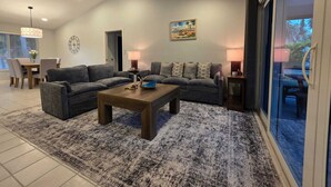 A living room with a gray sofa set, a wooden coffee table, and a patterned rug. There is a ceiling fan, a dining area in the background, and a large window with a view. - Pleasant 3-bedroom house with pool in peaceful setting (Naples)
