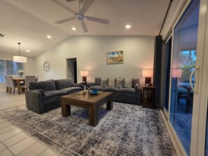 Ein Wohnzimmer mit einer grauen Sofagarnitur, einem Holztisch und einem gemusterten Teppich. Ein Deckenventilator, ein Essbereich im Hintergrund und ein großes Fenster mit Blick auf die Landschaft sind ebenfalls zu sehen.