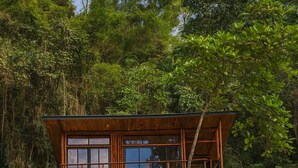 Una cabana de fusta de dues plantes amb grans finestres i un balcó, envoltada per un bosc dens.