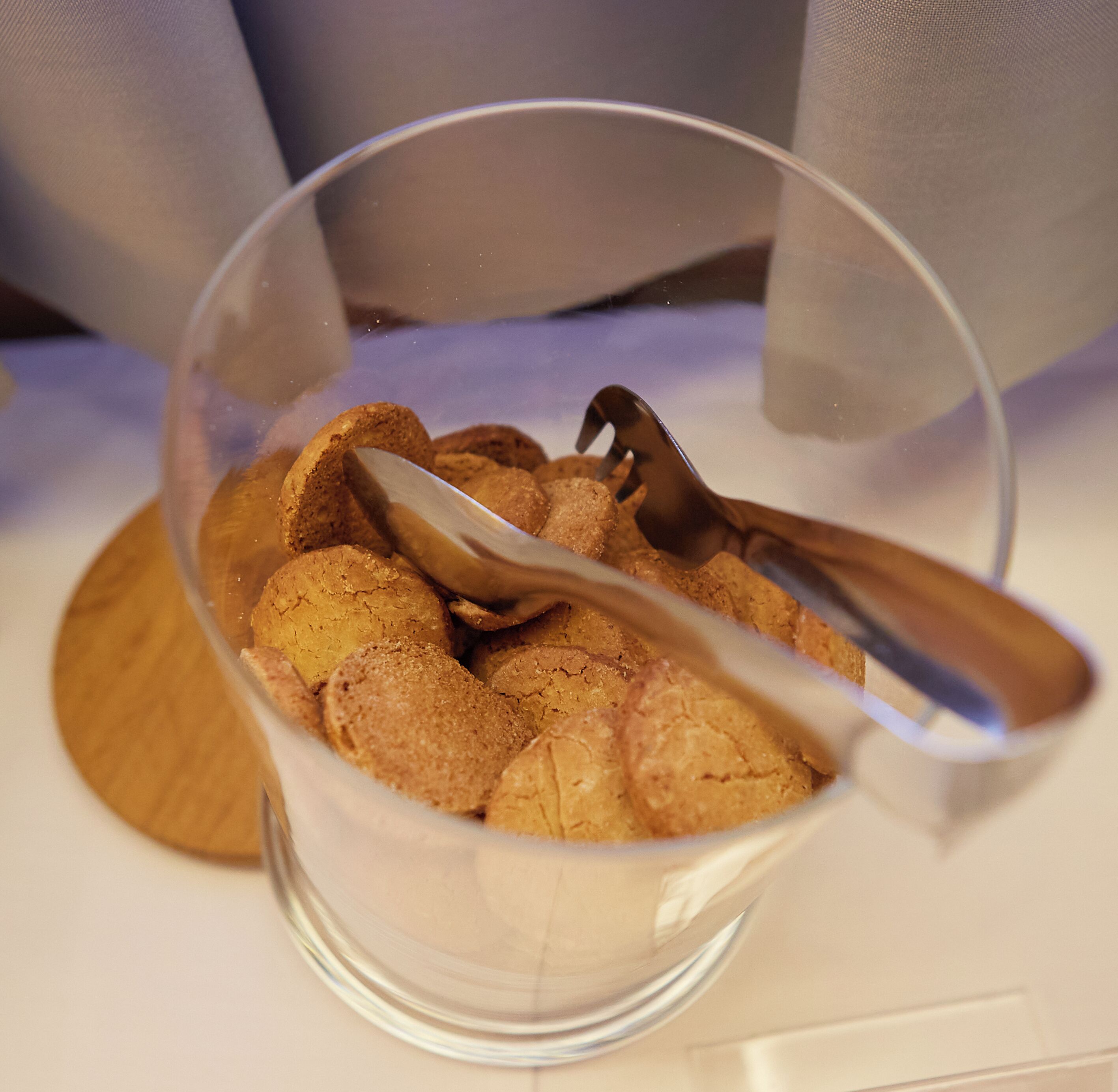 A clear glass bowl containing cookies with a spoon inside, placed on a wooden coaster.