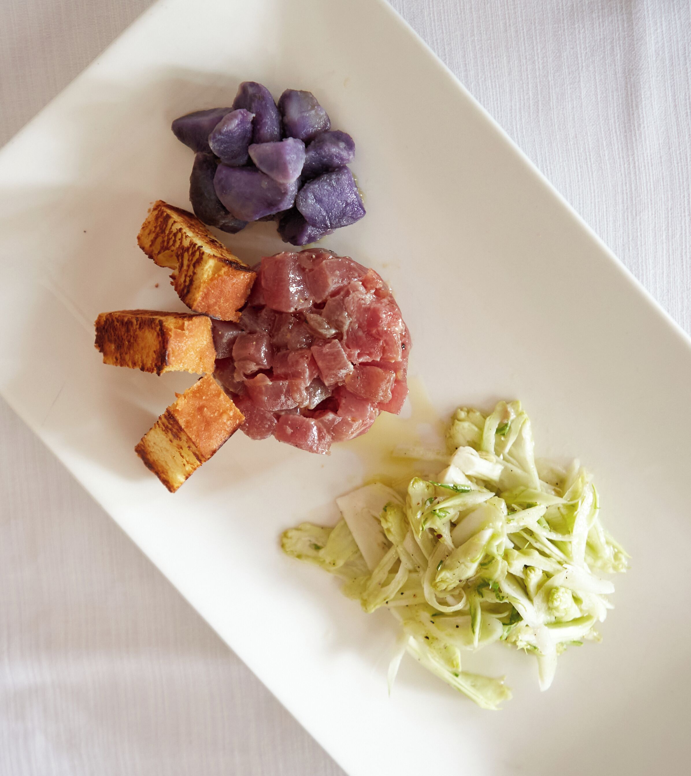 Ein weißer Teller mit verschiedenen Speisen, darunter violettes Gemüse, rohes Fleisch, frittierte Gerichte und gehobelte Krautsalat.