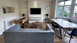 A modern living room with a sofa, a coffee table, a dining table with chairs, a foosball table, a TV mounted on the wall, and large windows with a view of houses outside. - Mayfield Escape - Stylish Two Bed Apartment (South Croydon)
