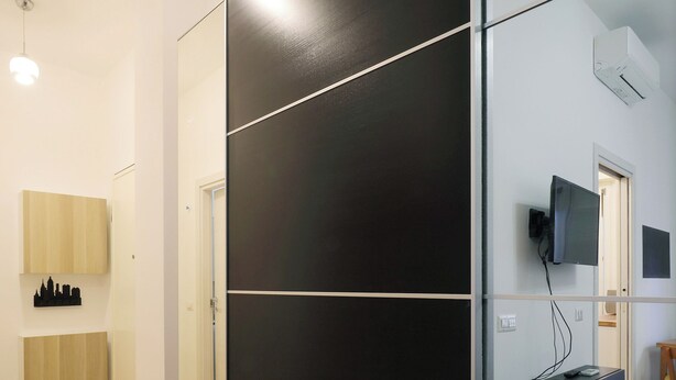 Una habitación de hotel moderna con suelo de madera, un panel divisor negro, un televisor de pantalla plana montado en la pared y una pequeña mesa de madera.