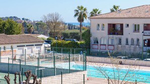 Un espace piscine avec un court de tennis entouré d’une clôture, un bâtiment doté de fenêtres en arc de cercle et une vue sur la mer.