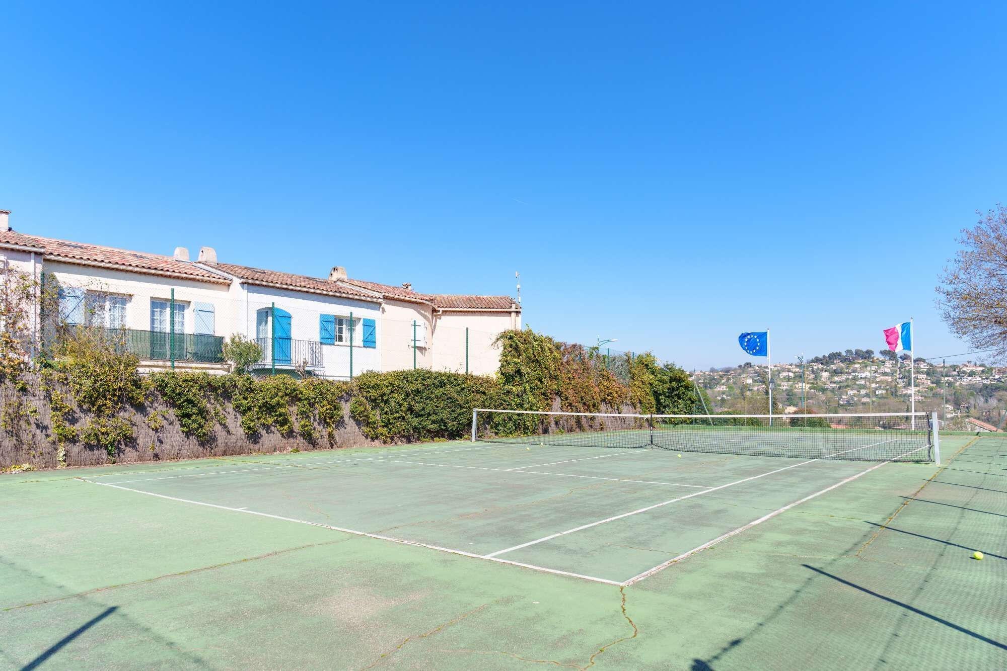 A tennis court with a net, surrounded by a white building with blue shutters, and flags in the background.