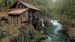 A rustic wooden house with a steeply pitched roof, situated on stilts over a flowing river, surrounded by dense forest. - Addendum USA (Detroit)