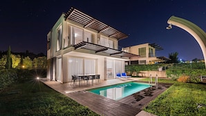 Una casa moderna de dos pisos con piscina, mobiliario exterior y un jardín bien iluminado en la noche.