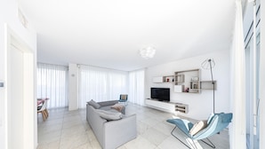 Una sala de estar moderna con un sofá gris, un sillón azul, un televisor de pantalla plana y una zona de comedor con mesa y sillas.